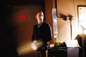 A man stands in a dimly lit room, holding a piece of paper. He is wearing a dark jacket over a checkered shirt and appears to be in deep thought or concentration. A microphone stand and various equipment are visible around him, hinting that this writer, perhaps an alumni like Jon Mooallem, is preparing for something significant.