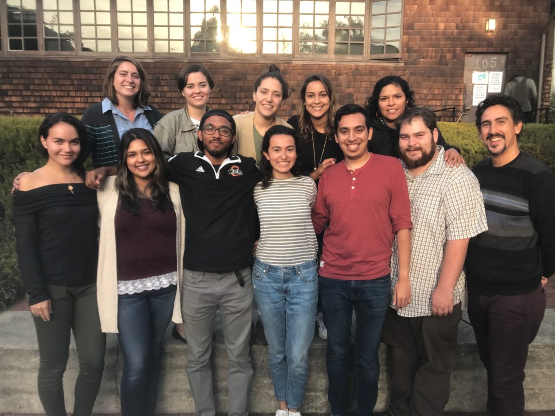 A group of twelve Hispanic journalists stands closely together, smiling at the camera. They are positioned in front of a building with wooden shingles and a large window. The people vary in age and style, wearing casual clothing, and display a sense of camaraderie.