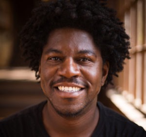 A person with short curly hair and a beard smiles warmly at the camera. They are wearing a dark-colored shirt, and the background is softly blurred, suggesting an indoor setting with light streaming in from the side. This is Serginho Roosblad, the esteemed Marlon T. Riggs Fellow.
