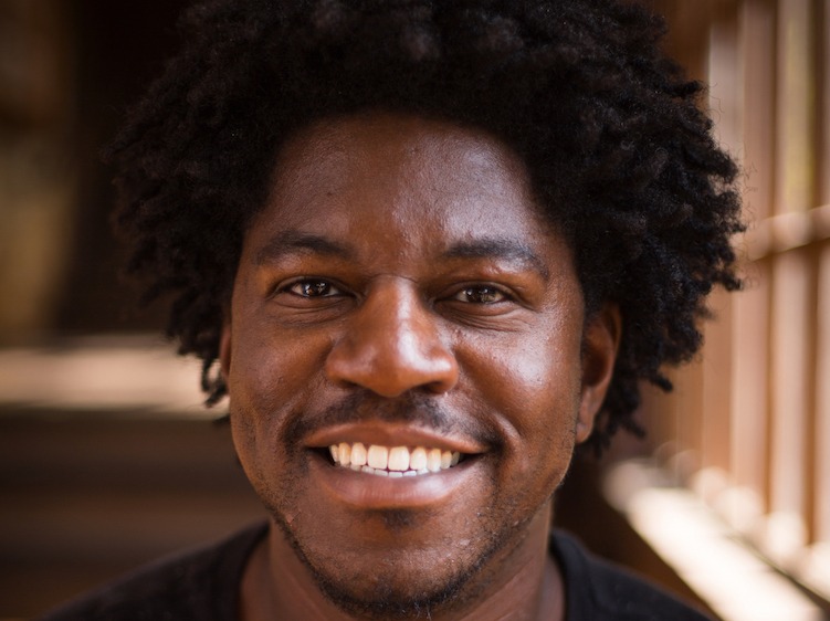 A man with curly hair and a beard, named Serginho Roosblad, smiles at the camera. He is wearing a black shirt, and the background appears to be a blurred indoor setting with wooden elements.