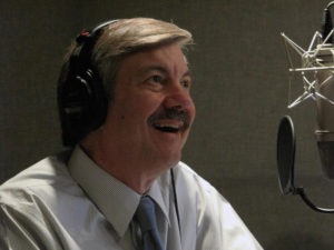 A man with a mustache, wearing headphones and a shirt with a tie, is smiling as he sits in front of a microphone. The background is a dimly lit room, reminiscent of a Berkeley Journalism recording studio.