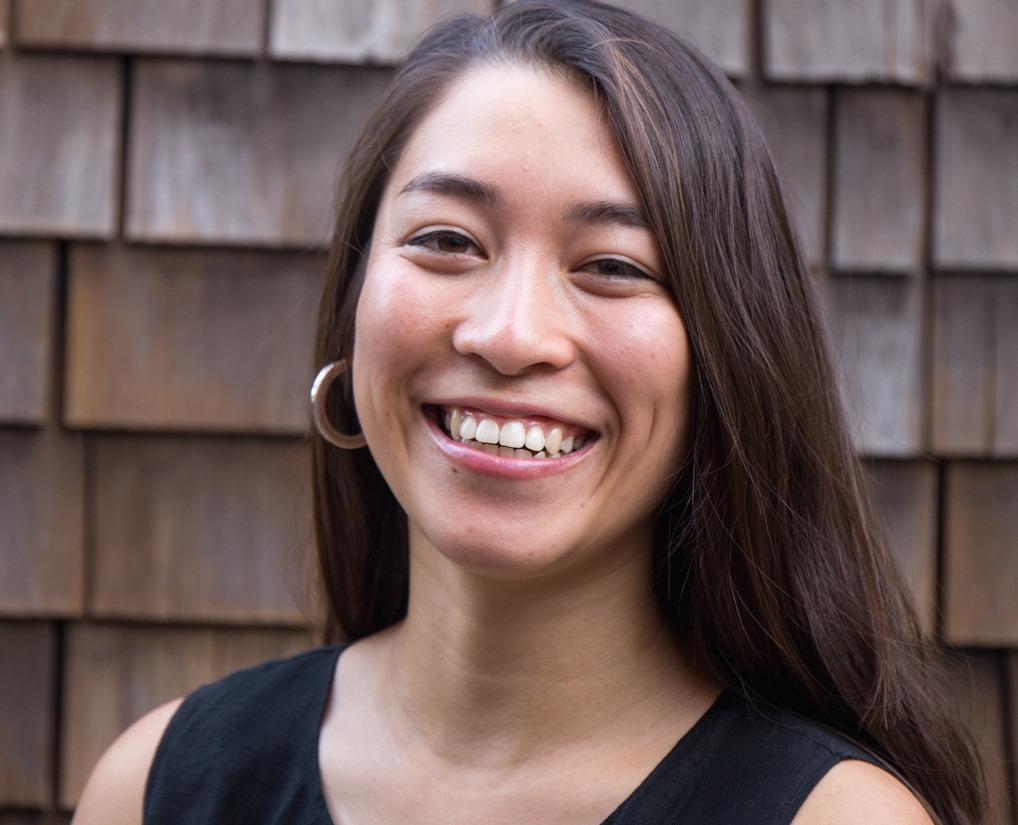 A person with long brown hair is smiling broadly at the camera. They are wearing a sleeveless black top and hoop earrings. The background consists of brown wooden shingles, reminiscent of the rustic charm often found near Berkeley Journalism school.