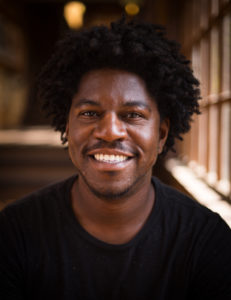 A man with curly hair and a beard smiles warmly at the camera. He is wearing a black t-shirt and is pictured indoors with a blurred background featuring wooden elements and natural light streaming in from a window, capturing the essence of Berkeley Journalism.