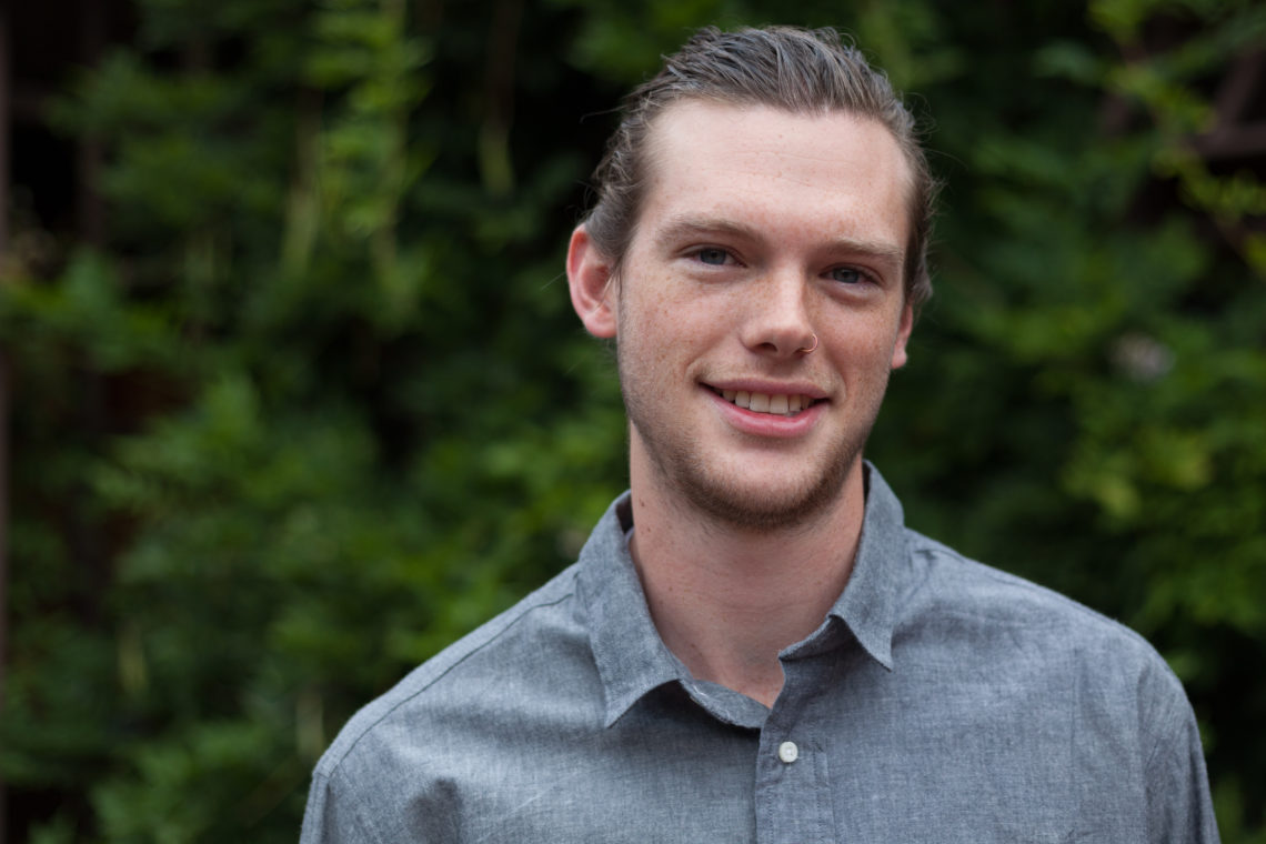 A young man with light skin, short hair pulled back, and a sparse beard smiles at the camera. Wearing a gray button-up shirt, he stands against a backdrop of lush green foliage. His confident look hints at his Berkeley Journalism aspirations.