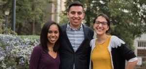 Three people are standing outdoors, smiling at the camera. The person on the left is wearing a dark purple long-sleeved shirt, the person in the middle is wearing a suit jacket over a striped shirt, and the person on the right is wearing glasses and a yellow top. Trees and buildings, including Berkeley Journalism's campus, are in the background.