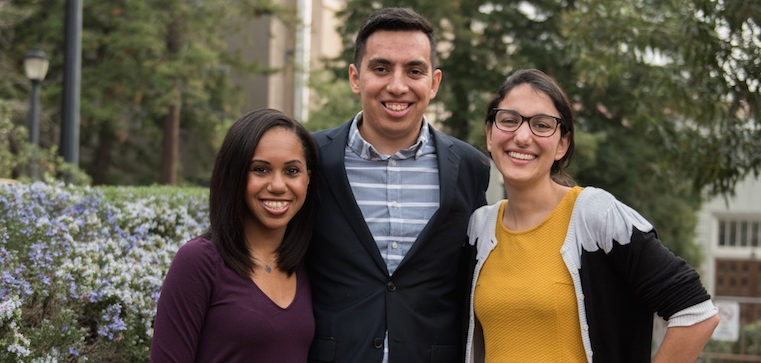 Three people are standing outdoors, smiling at the camera. The person on the left is wearing a dark purple long-sleeved shirt, the person in the middle is wearing a suit jacket over a striped shirt, and the person on the right is wearing glasses and a yellow top. Trees and buildings, including Berkeley Journalism's campus, are in the background.