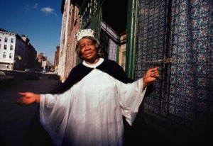 An elderly person stands outside a building on a sunny day, wearing a white dress, a dark cape, and a tiara. The person is smiling and gesturing with one hand. Behind them is a patterned wall as the city street stretches out to the left—a scene reminiscent of Berkeley Journalism's unique storytelling approach.