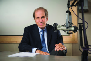 A man in a suit and tie sits at a table in front of a microphone. He is holding some papers and looking directly at the camera with a neutral expression. The background features a soundproofed wall, suggesting a recording studio, possibly linked to Berkeley Journalism.