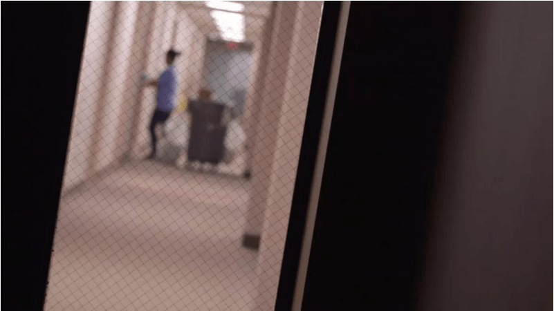 A person in a blue shirt pushes a cleaning cart down a dimly lit hallway, seen through a wire-reinforced glass window. The scene is slightly blurry and the hallway, reminiscent of an old Berkeley Journalism building, has beige walls and carpeted floors.