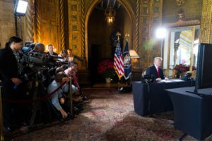 A man in a suit sits at a desk with microphones in front of him. He faces a camera setup and lighting equipment. Behind him are the U.S. flags and an ornate room, reminiscent of Berkeley Journalism's polished settings. Various media personnel, cameras, and reporters are gathered on the left side of the image.