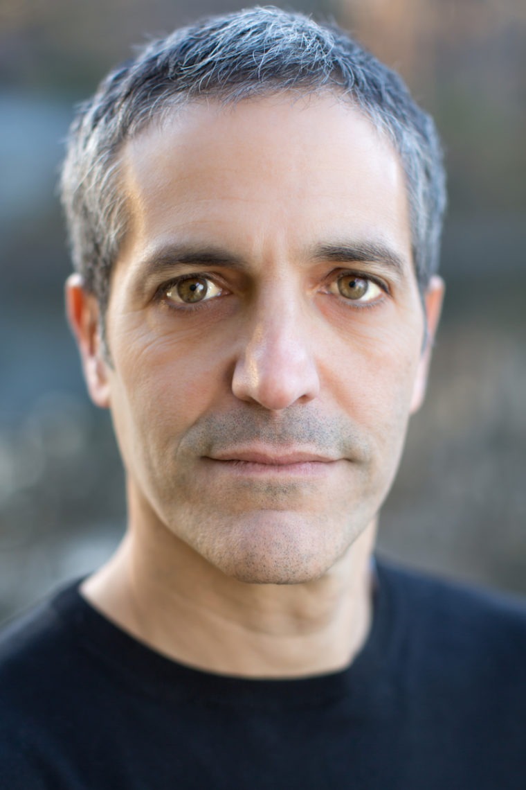 A middle-aged man with short graying hair and brown eyes looks directly at the camera. He has a calm expression, framed by a plain, slightly blurred background. Wearing a black shirt, he exudes the understated confidence often seen in Berkeley Journalism professionals.