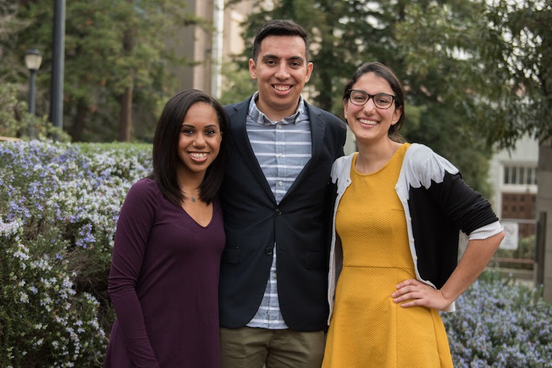 Three people standing outdoors, smiling. From left to right: a person in a maroon long-sleeve top, a person in a black jacket over a striped shirt, and a person in a mustard yellow dress with a black and white cardigan. They are standing in front of flowering bushes and trees on the Berkeley Journalism campus.