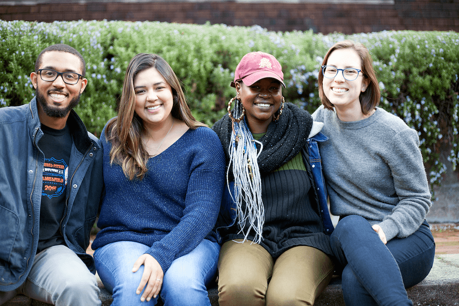 A group of four people, connected through Berkeley Journalism, sitting outdoors and smiling at the camera. They are dressed in casual clothing with lush greenery visible behind them.