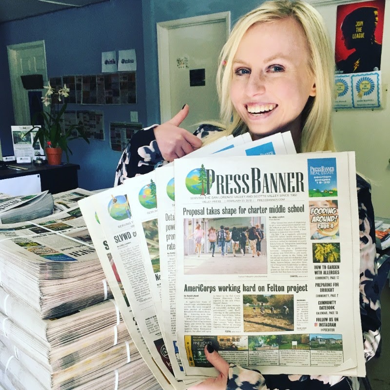 A smiling person with blonde hair, wearing a light green shirt, holds up several copies of the Press Banner newspaper inside a brightly lit office. The office, reminiscent of Berkeley Journalism vibes, has plants, stacks of newspapers, and posters on the walls.