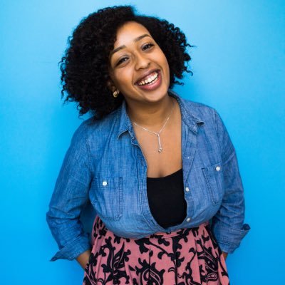 A person with curly hair smiles brightly against a vibrant blue background. They are wearing a denim shirt over a black top and a pink patterned skirt, accessorized with a silver necklace. Their hands are placed in the pockets of their skirt, embodying the eclectic style often seen around Berkeley Journalism.