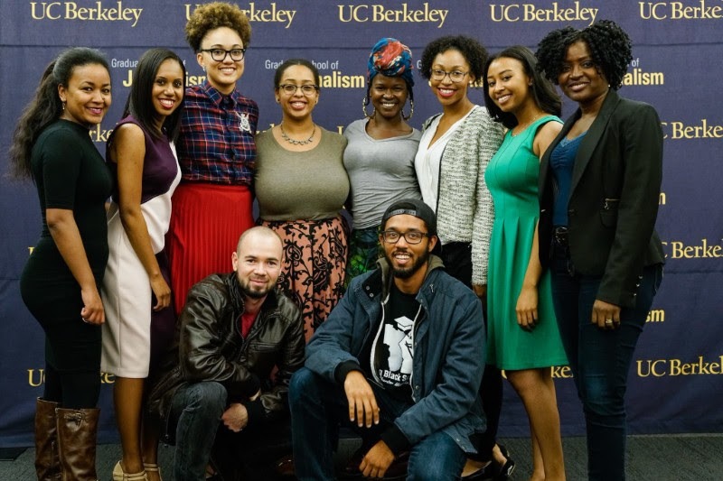 A diverse group of nine people, eight standing and one kneeling in front, smile for a photo in front of a UC Berkeley Journalism backdrop. The group includes both men and women, all dressed in various styles of casual attire. The background reflects affiliation with UC Berkeley.