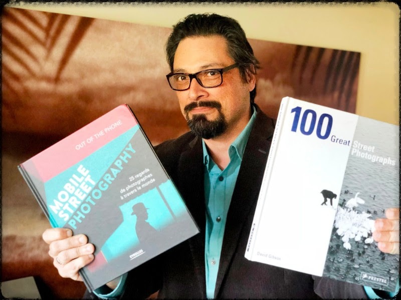 A bearded man with glasses, dressed in a teal shirt, is holding two photography books. In his right hand is 