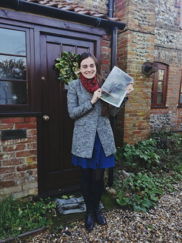 A person is standing in front of a wooden door with a wreath, holding a printed photograph. Dressed in a gray coat, red scarf, blue dress, and black tights, they exude the thoughtful presence of someone from Berkeley Journalism. The door belongs to a brick and stone building with a garden path made of gravel.