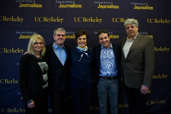 Five people are standing together, smiling, with their arms around each other in front of a backdrop that reads 