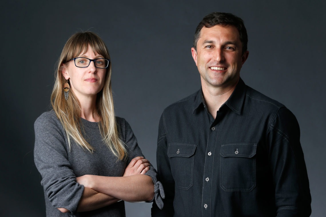 A woman with long blonde hair, wearing glasses and a gray sweater, stands with her arms crossed beside a smiling man with short dark hair wearing a black button-up shirt. They are posed in front of a dark, neutral background, embodying the professionalism typical of Berkeley Journalism.