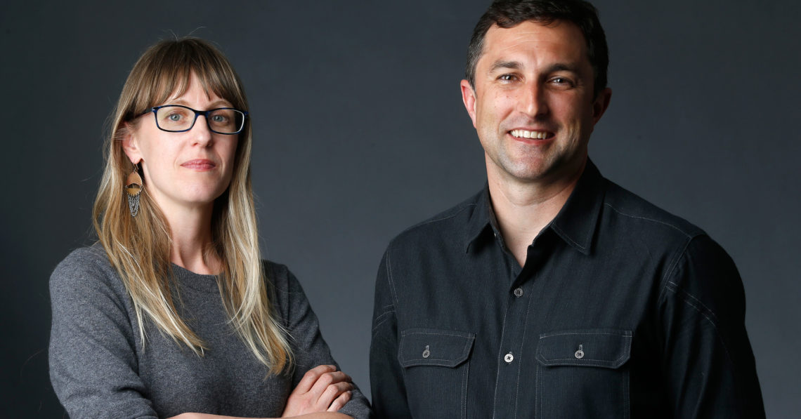 A woman with long blonde hair, wearing glasses and a grey sweater, stands beside a man with short dark hair, wearing a black collared shirt. Both are standing against a dark background and looking at the camera, with the man smiling broadly. They seem ready for an engaging Berkeley Journalism project.