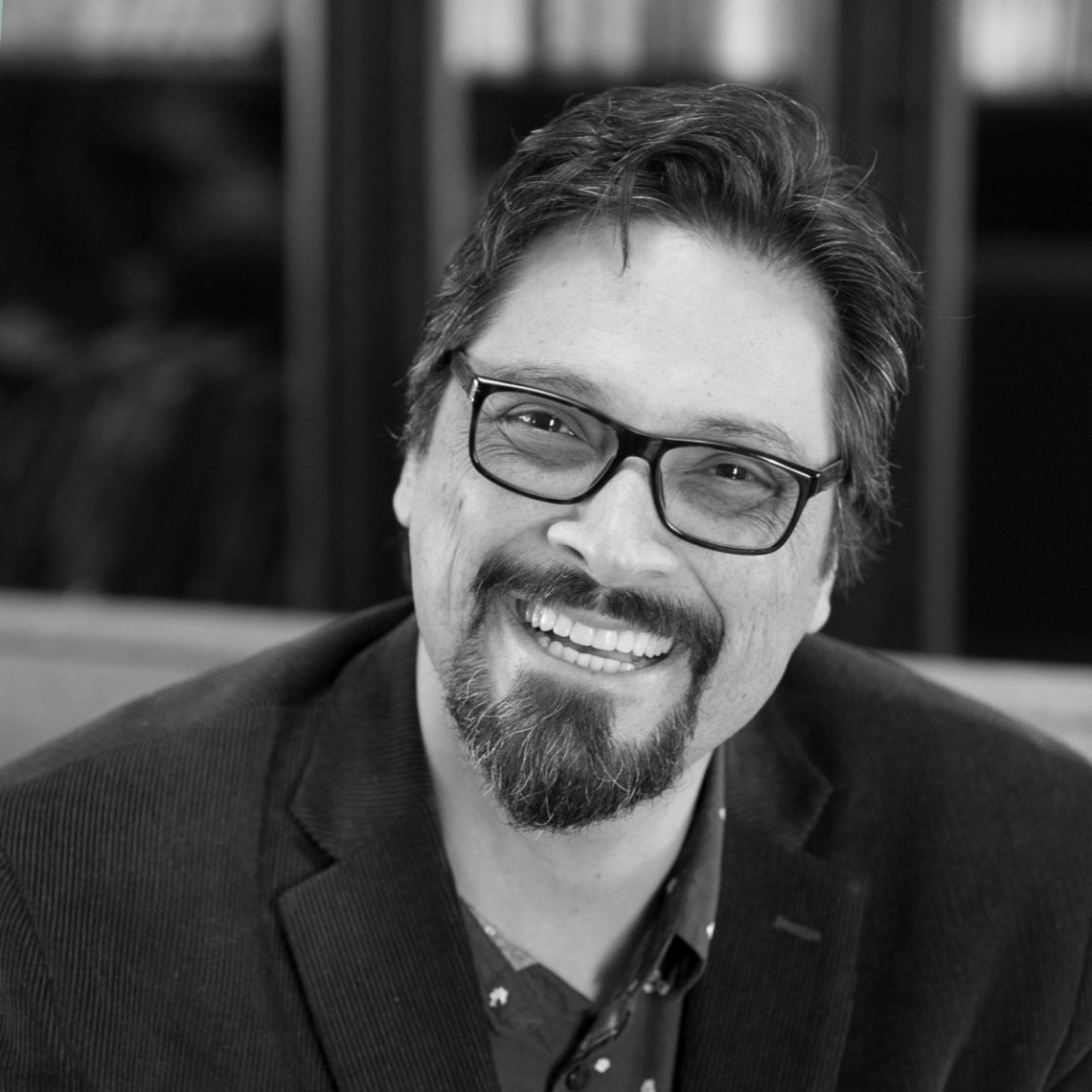 A man with glasses and a beard smiles warmly at the camera. He is wearing a dark blazer over a patterned shirt. The background is slightly blurred, focusing attention on his face. This black-and-white photograph captures an approachable expression, embodying the spirit of Berkeley Journalism.