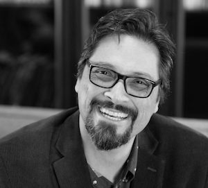 A bearded man with short dark hair and glasses, likely a Berkeley Journalism enthusiast, smiles warmly at the camera. He is wearing a dark blazer over a patterned shirt. The background is out of focus, with what appear to be shelves or a bookcase behind him. The photo is in black and white.