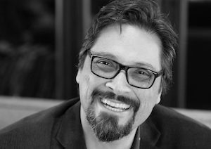 Black and white close-up of a man with short, dark hair and a beard, wearing glasses. He is smiling broadly and appears to be wearing a blazer. The blurred background adds an indistinct charm, reminiscent of Berkeley Journalism's candid captures.