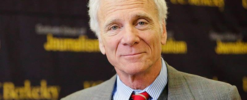 An older man with white hair and a kindly expression is wearing a gray suit, blue shirt, and red tie. He stands in front of a backdrop with repeated text that reads "Berkeley Journalism.
