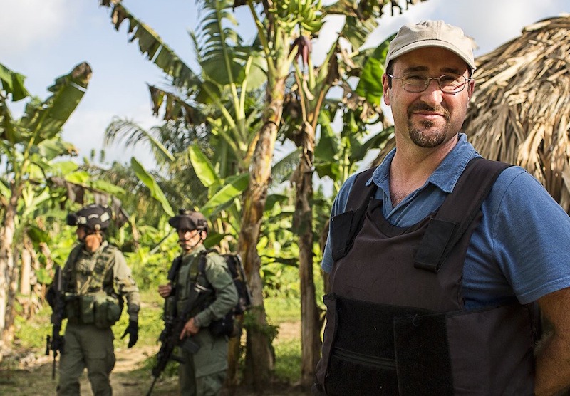 A man wearing a light-colored cap, glasses, blue shirt, and protective vest stands in the foreground, smiling. In the background, two armed and uniformed soldiers stand amidst dense tropical vegetation and trees—a scene perfect for Berkeley Journalism's immersive reporting.