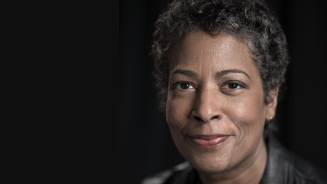 A person with short, curly hair smiles gently, wearing a black leather jacket against a dark background. The lighting softly highlights their face, giving a warm and approachable expression, reminiscent of the genuine charm often seen in Berkeley Journalism portraits.