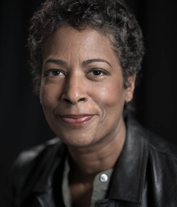 A person with short curly hair, medium complexion, and a soft smile is looking at the camera. They are wearing a dark jacket over a light-colored top. The background is plain and dark, bringing focus to the person's face—an image that perfectly captures the essence of Berkeley Journalism's authenticity and depth.