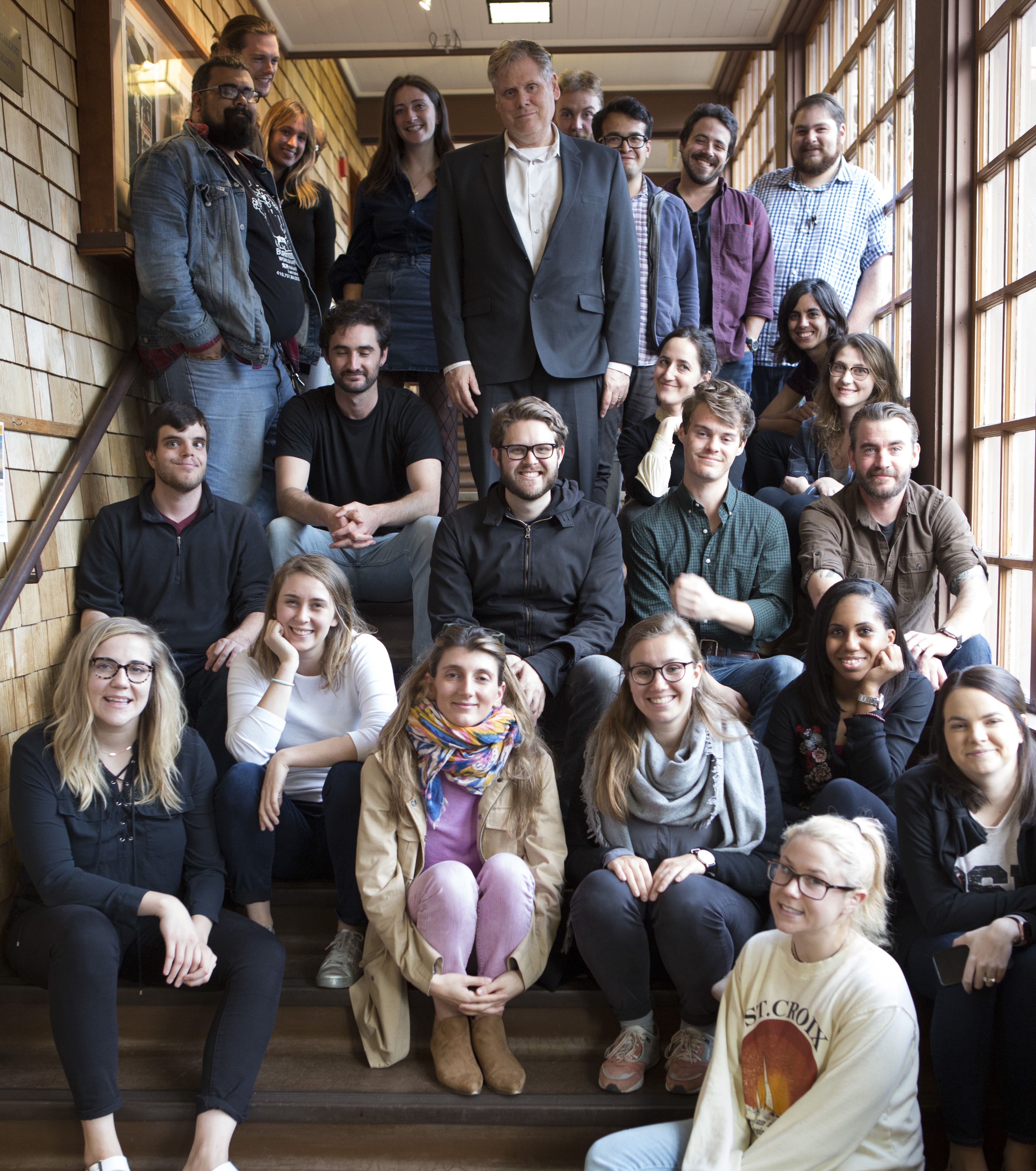 A group of 25 people, including several Berkeley Journalism students, pose on a staircase inside a building with wooden walls. Some are sitting on steps, while others stand behind them. They appear to be a mix of men and women in casual outfits. Many are smiling, and the mood looks cheerful and friendly.