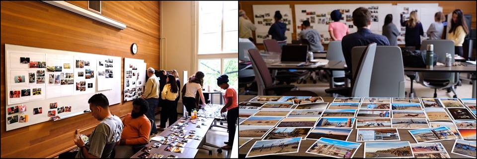 A group of people in a classroom-like setting are engaged in an activity involving photos, similar to those found at Berkeley Journalism. One part of the room has several photos displayed on boards, with some standing and examining them. On the right, people are seated, with a table full of photos in the foreground.