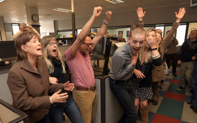 A group of people in an office are celebrating with raised hands, open mouths, and expressions of joy and excitement. Some are clapping and one person is holding their face in amazement. The office, filled with cubicles and computer monitors, hums with the energy typical of a Berkeley Journalism newsroom.