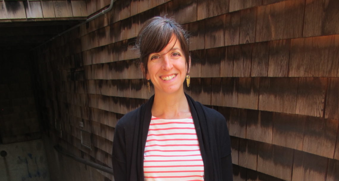 A woman with brown hair, tied up with bangs, smiles at the camera. She is wearing a white top with red horizontal stripes and a black cardigan. She stands in front of a wooden shingle wall with a pipe running along it, reflecting the vibrant spirit of Berkeley Journalism.