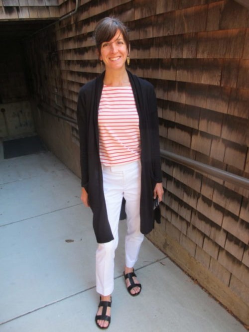 A person stands outside, smiling, on a concrete pathway next to a building with wooden shingle siding. They are wearing a red and white striped top, white pants, a black cardigan, and black sandals. Their hair is tied back, and they seem content and relaxed—perhaps on their way to Berkeley Journalism.