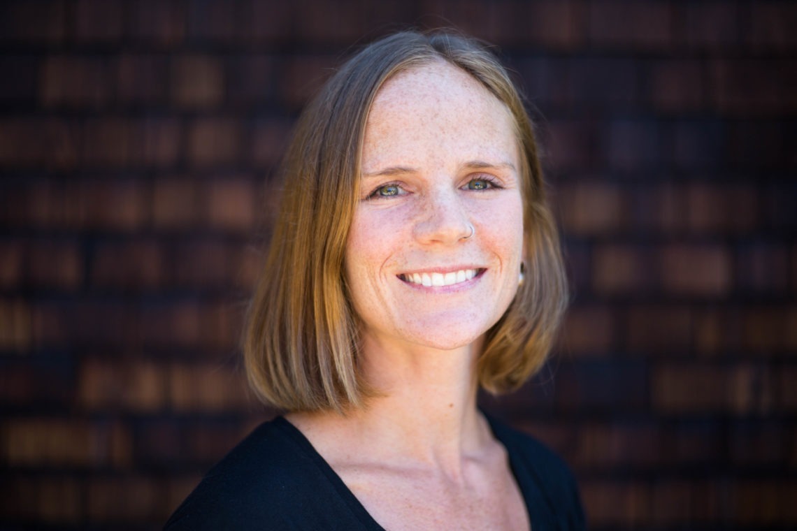 A woman with shoulder-length brown hair and light skin smiles at the camera. She has freckles on her face and is wearing a black top. The background consists of a dark, blurry wooden surface, setting the scene for an inspiring Berkeley Journalism portrait.