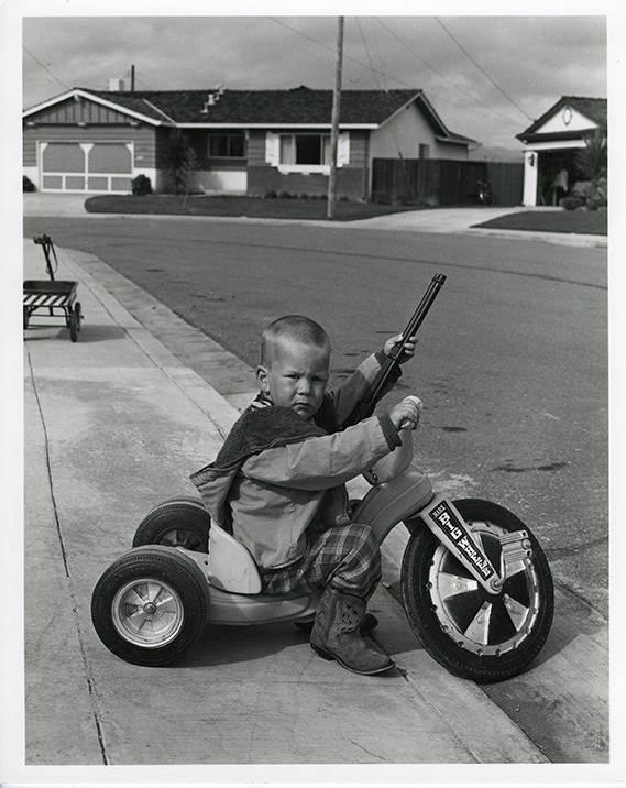 A young child with a serious expression rides a tricycle on the sidewalk in a suburban neighborhood. Dressed in a jacket and boots, they hold a toy rifle. Houses with lawns and driveways are visible in the background, painting a scene that looks like it emerged from Berkeley Journalism archives.