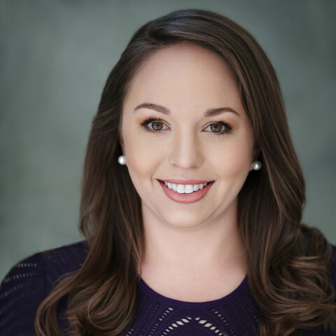 Katey Rusch, a smiling woman with long brown hair, wears a purple top and pearl earrings against a neutral background.