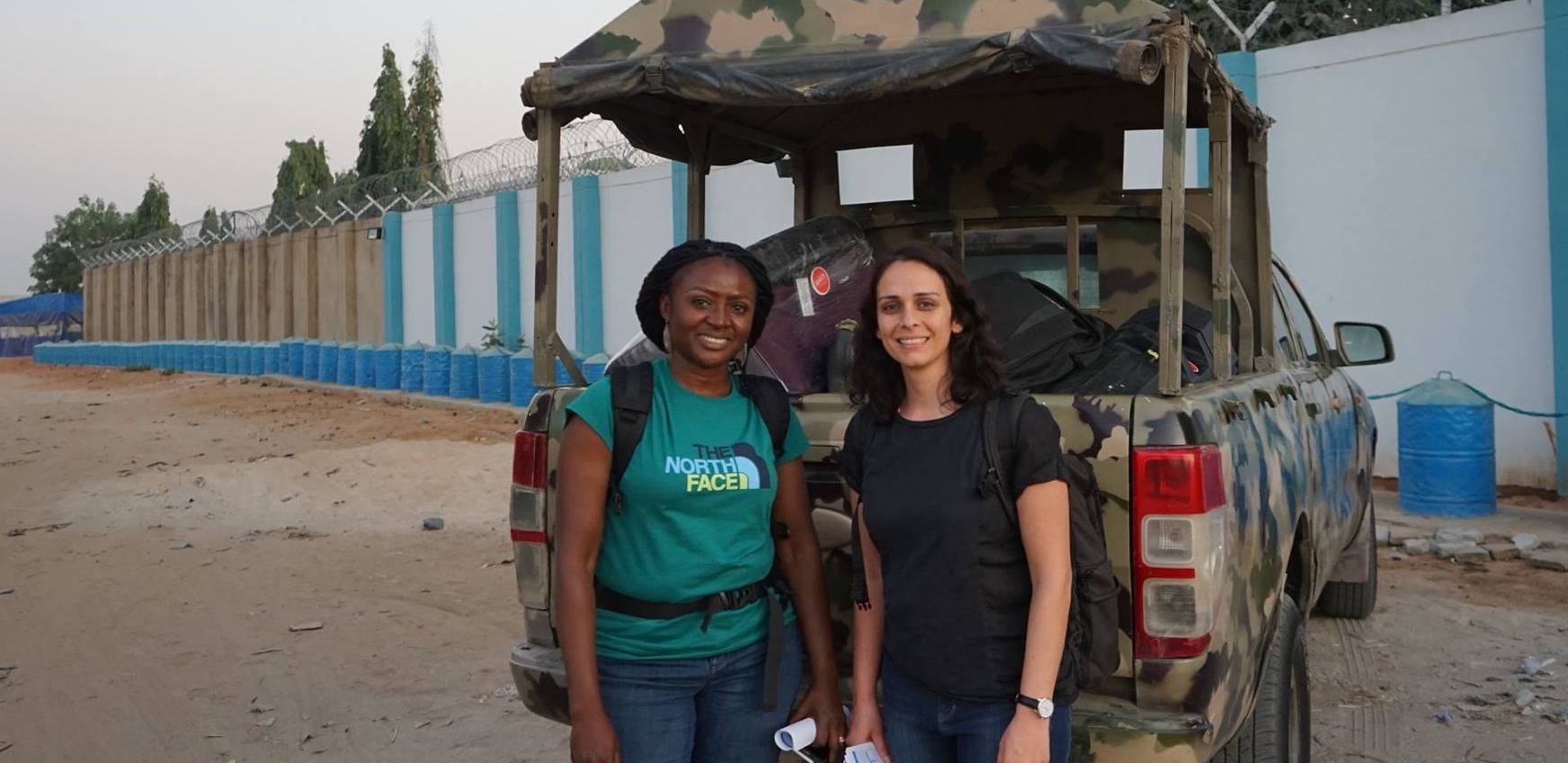 Two women stand in front of a camouflage-painted pickup truck. One, wearing a green shirt and the other wearing a plain black shirt.