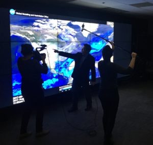 A person stands in front of a large digital map display, pointing at the screen, while speaking to a Berkeley Journalism camera crew consisting of a videographer and a sound technician with a boom microphone. The display shows a world map with weather patterns and data overlays.