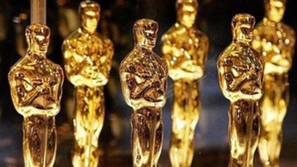 A close-up image of several golden Oscar statuettes standing in rows. The statuettes have a human figure design with arms crossed over their chest, mounted on a black base, and are highly reflective under the light. The background is out of focus, reminiscent of the prestigious awards often covered by Berkeley Journalism.