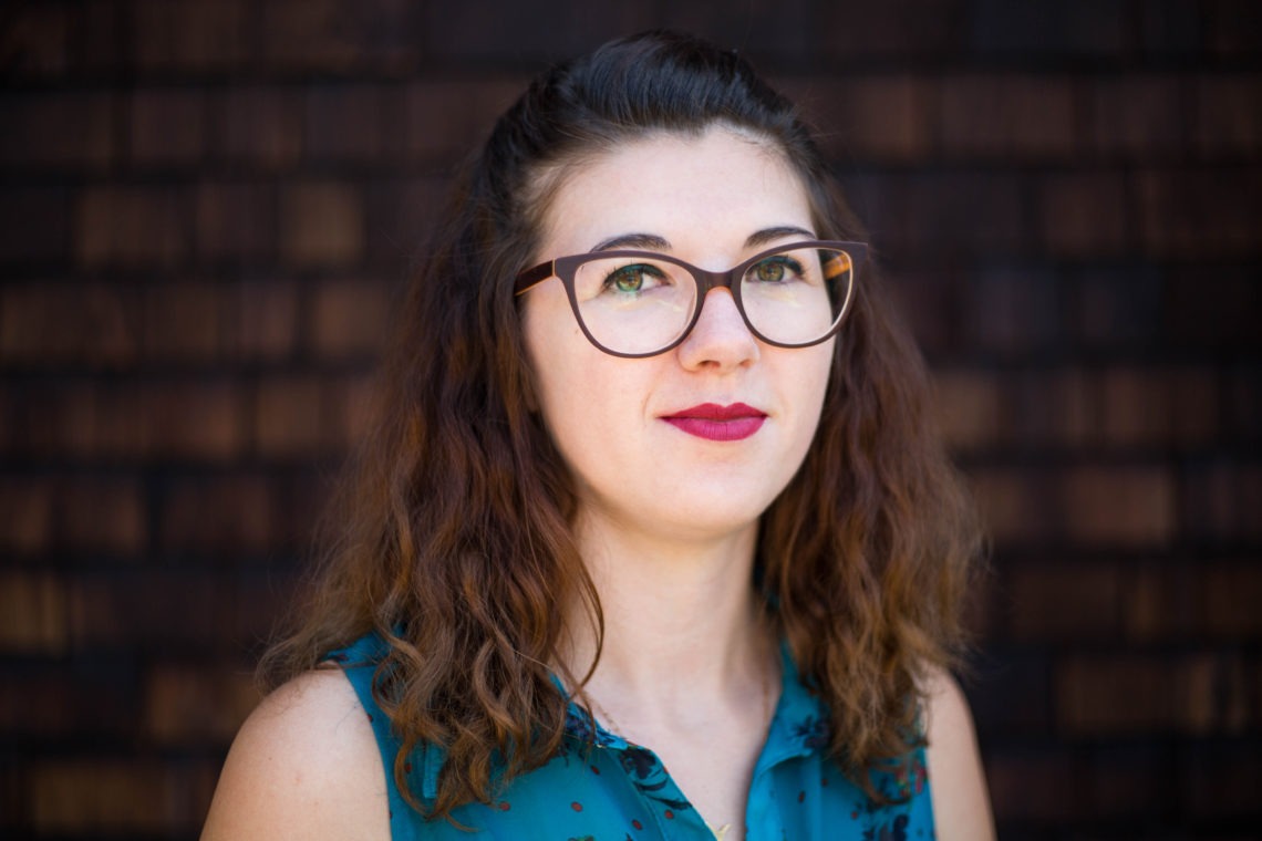 A woman with wavy brown hair and glasses is standing in front of a dark wooden background. She is wearing a sleeveless green top with a subtle pattern and has a slight smile on her face, embodying the confident and inquisitive spirit of Berkeley Journalism.