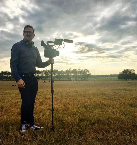 A young man holding camera equipment outside on a farm. The sky is cloudy with sun peaking through. 