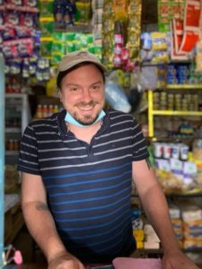 A man with a beard and mustache, likely John Fecile, is wearing a black and blue striped shirt, cap, and face mask around his neck. He's smiling while standing behind a counter in a store filled with various colorful packages and products on shelves.