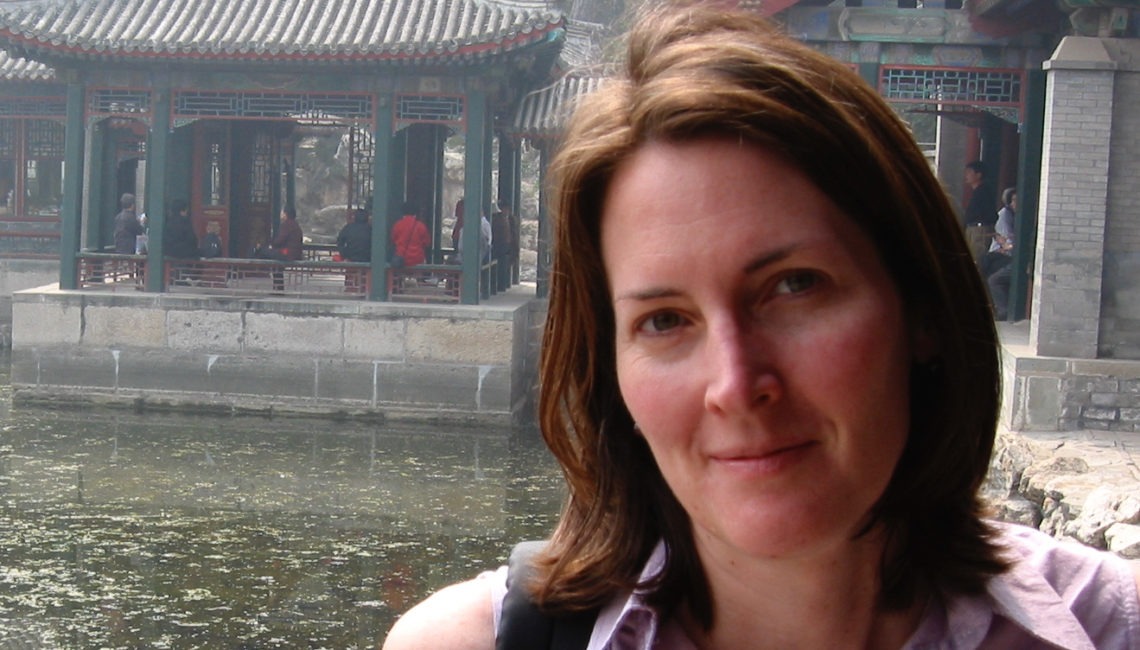 A woman with shoulder-length brown hair stands in front of traditional Chinese architecture, including buildings with tiled roofs and intricate wooden details. A pond with murky water can be seen in the foreground. She is smiling slightly and wearing a sleeveless top, capturing the serene moment for Berkeley Journalism.