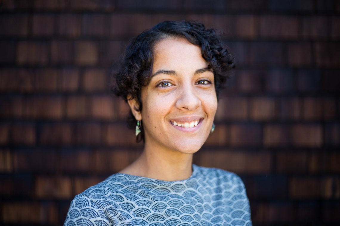 A smiling person with short, curly hair is wearing a patterned shirt and small earrings, reflecting the stylish flair often seen at Berkeley Journalism. The background consists of dark brown, wooden shingles.