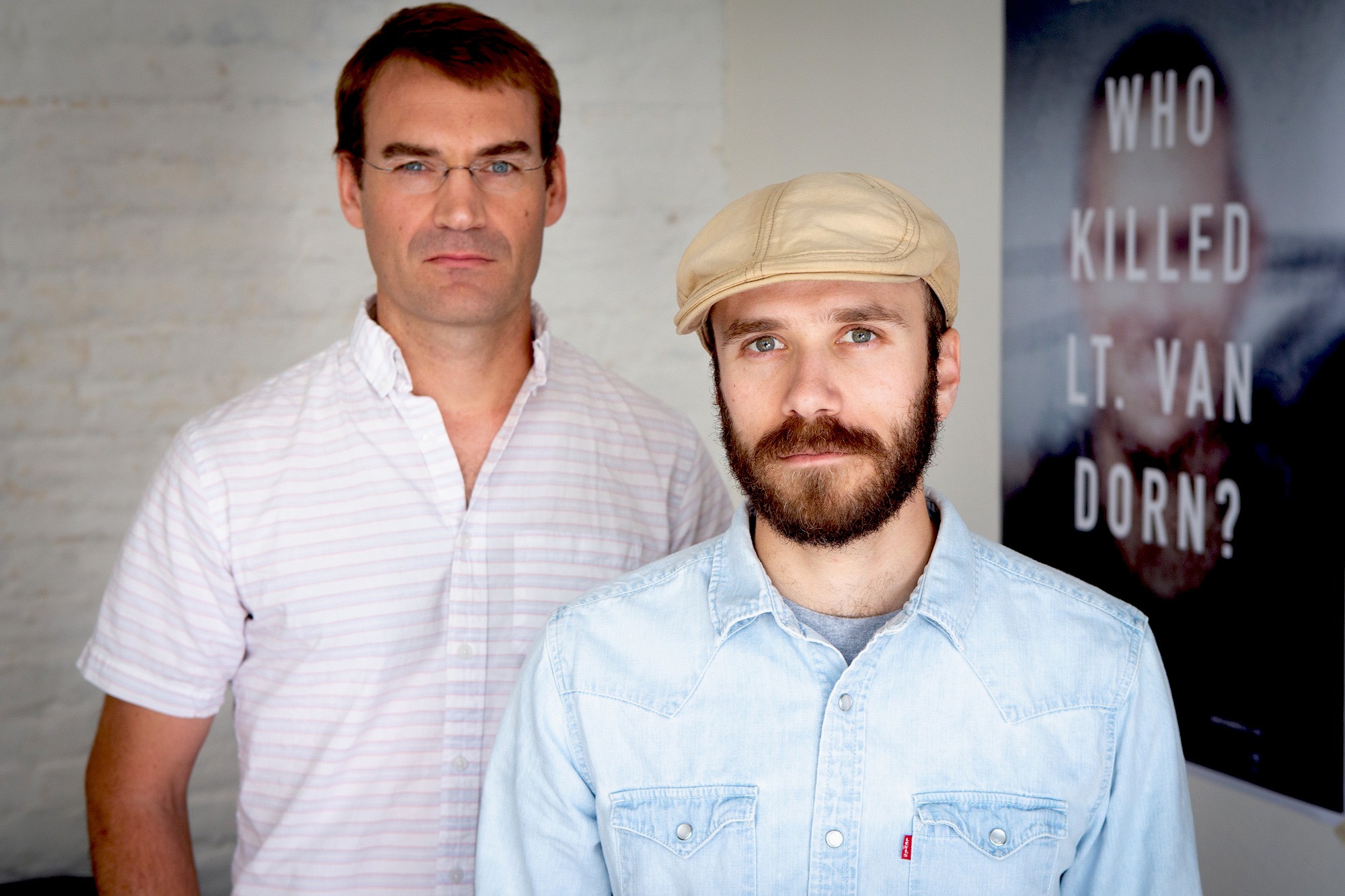 Two men are standing indoors, looking directly at the camera. The man on the left is wearing glasses and a light-colored striped shirt. The man on the right is wearing a beige cap, a beard, and a light denim shirt. A poster on the wall behind them reads 