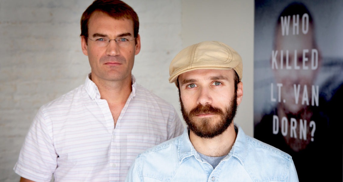 Two men stand indoors, the man on the left wearing glasses and a light-colored button-down shirt, and the man on the right wearing a beige cap and a light blue shirt. A blurred poster in the background reads "WHO KILLED LT. VAN DORN?". It seems like a scene straight out of Berkeley Journalism.